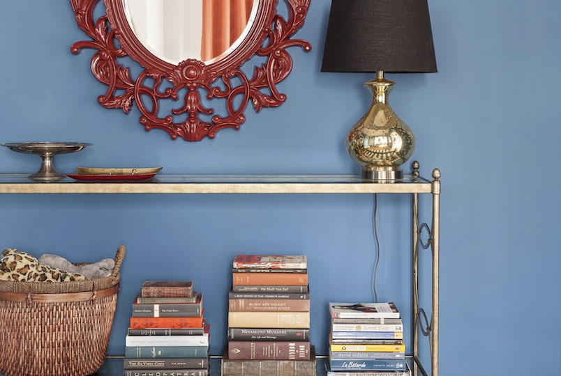 interior designed room : shelf of books and basket, red ornate framed mirror, short brass lamp with black shade