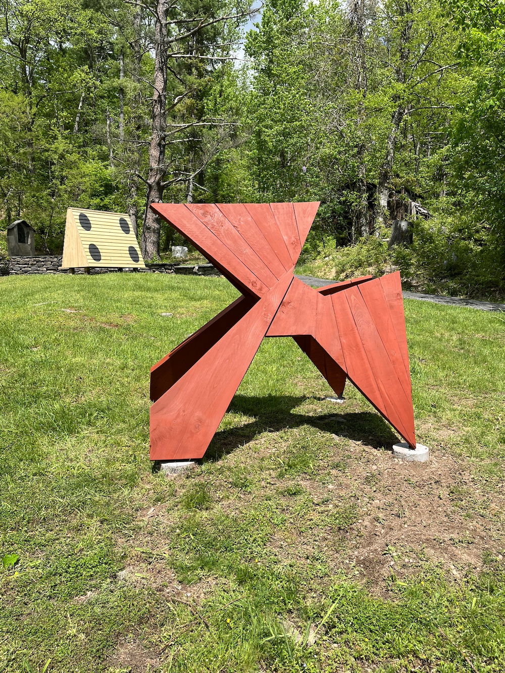red painted axel shaped sculpture in the grass with another yellow sculpture in the distance