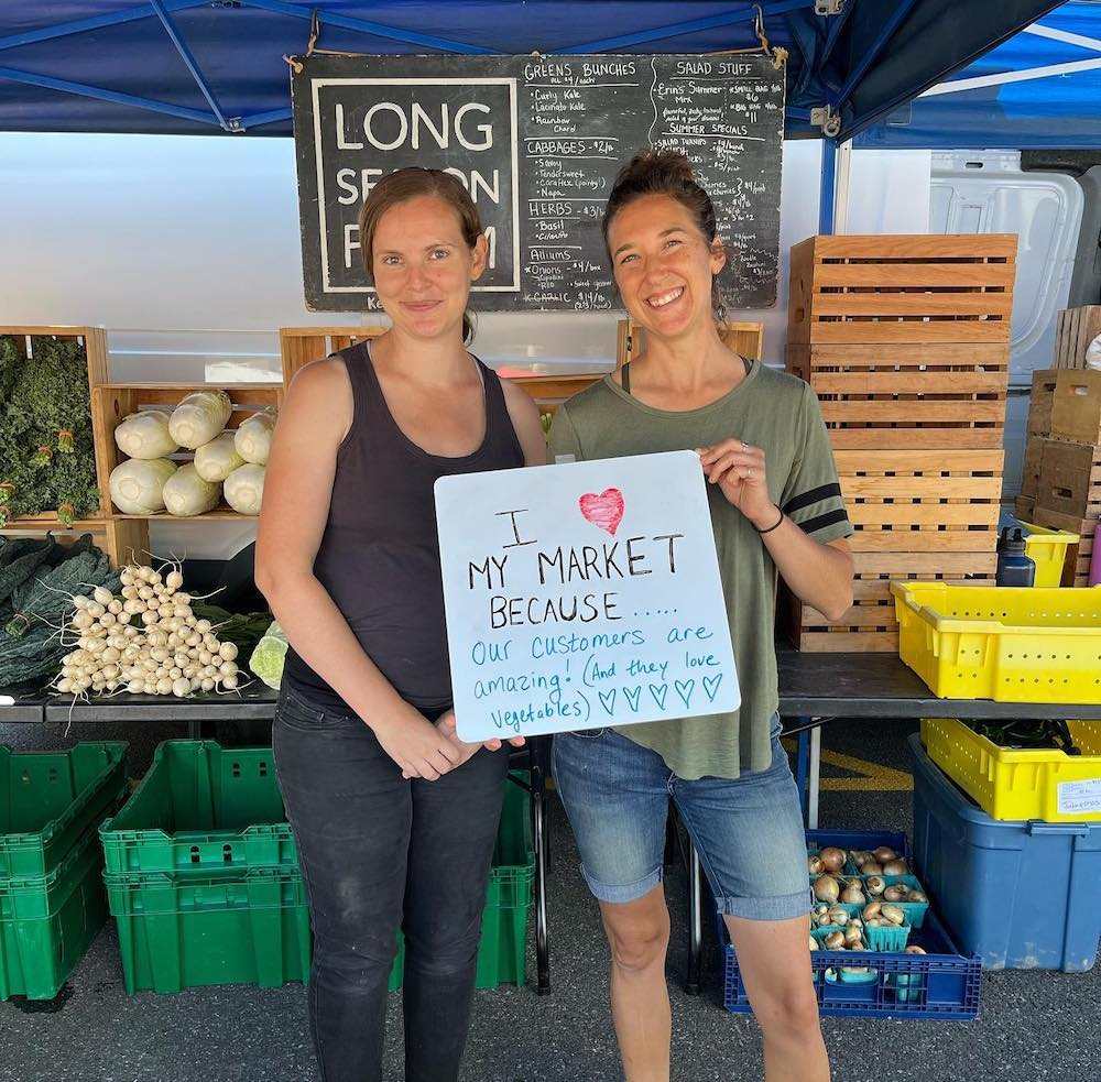 two women holding a sign about why they love farmers markets