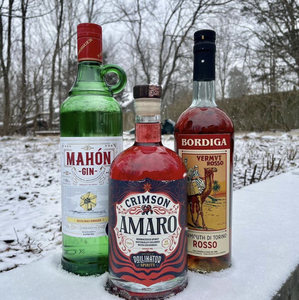 three bottles of spirits to be used to make negronis on a fence with a winter scene in the background to buy at Woodstock Wine & Liquors in Woodstock, NY